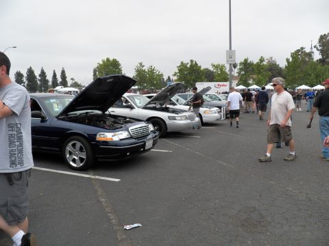 2003 Ford Crown Victoria - blue sedan with hood up leads a line of cars at a show; people mill about near a Ryder banner.