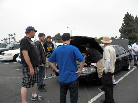 2003 Ford Crown Victoria - Spectators surround a black sedan as a man works under its raised hood in a sunny parking lot.