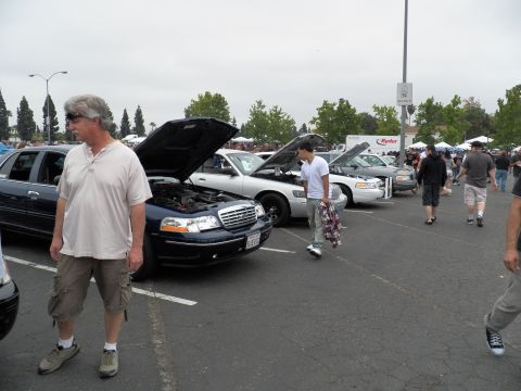 2003 Ford Crown Victoria - man in cargo shorts strolls past a blue Ford sedan with its hood raised among other show cars.