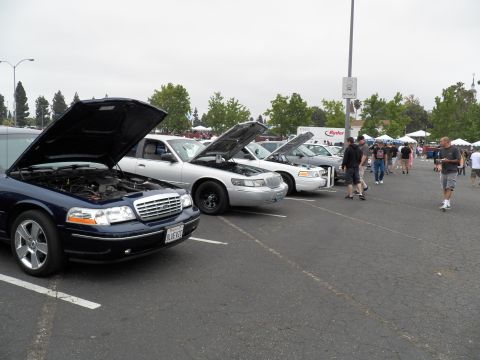 2003 Ford Crown Victoria - Blue Ford sedan with open hood leads a row of cars; attendees walk the lot under a gray sky.