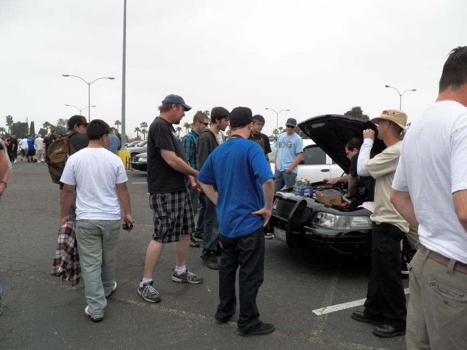 Crowd of men around white sedan with open hood at outdoor parking-lot car show