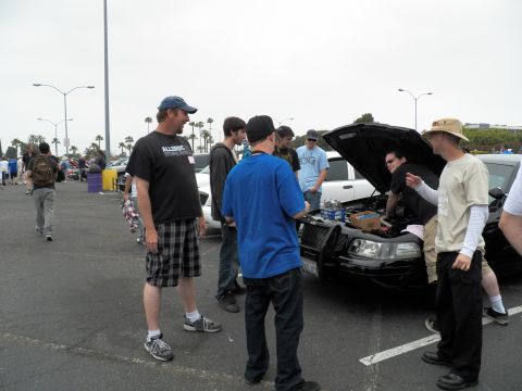 2003 Ford Crown Victoria - group converses around a white sedan's open hood, bottled water on the engine, parking lot behind.