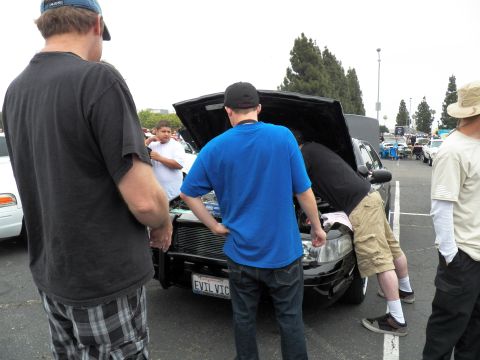 2003 Ford Crown Victoria - Onlookers inspect the engine bay of a black sedan displaying an EVIL VIC California plate at a parking-lot car show.