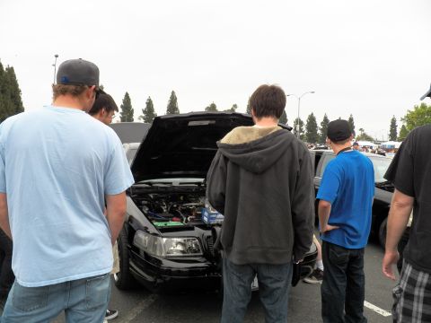 2003 Ford Crown Victoria - Several attendees stand around the open hood of a dark sedan among rows of cars at a meet.