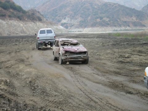1985 Ford Bronco - Two vehicles, one heavily damaged, travel on a muddy dirt road through a barren landscape.