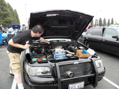 2003 Ford Crown Victoria - man leans into the engine bay of a black sedan with front push bar and EVIL VIC plate at a show.