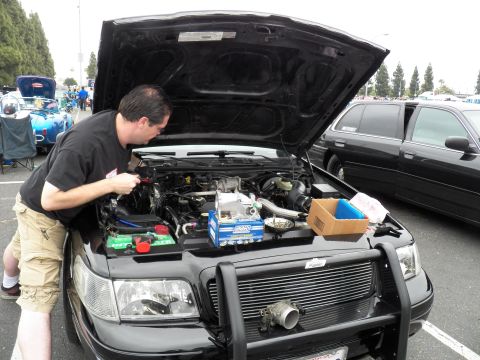 2003 Ford Crown Victoria - man in a black shirt and tan shorts works under the raised hood of a black sedan with a brush guard.