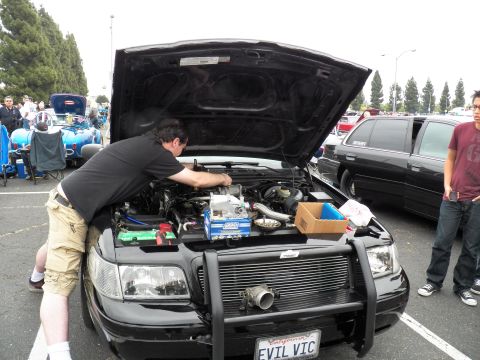 2003 Ford Crown Victoria - man leans across the engine of a black sedan marked EVIL VIC as a bystander watches at the show.