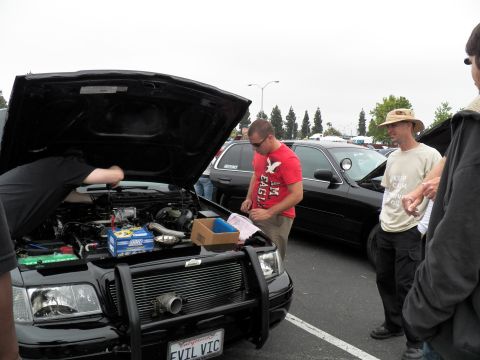 2003 Ford Crown Victoria - man in a red shirt reads a flyer beside a black sedan with raised hood and EVIL VIC plate.