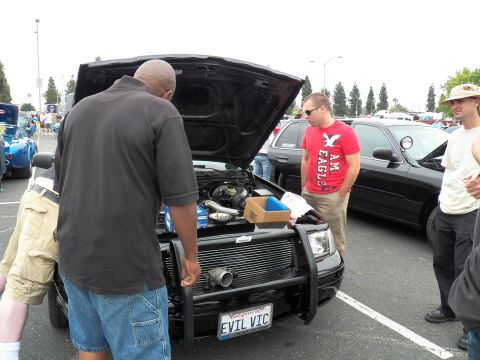 2003 Ford Crown Victoria - Attendees gather at the open engine bay of a black sedan displaying an EVIL VIC California plate.
