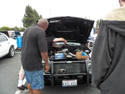 2003 Ford Crown Victoria - man works under the hood of a black sedan with grille guard and EVIL VIC plate while others watch.