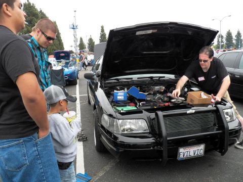 2003 Ford Crown Victoria - child and adults watch a man work on the engine of a black sedan marked EVIL VIC at a show.