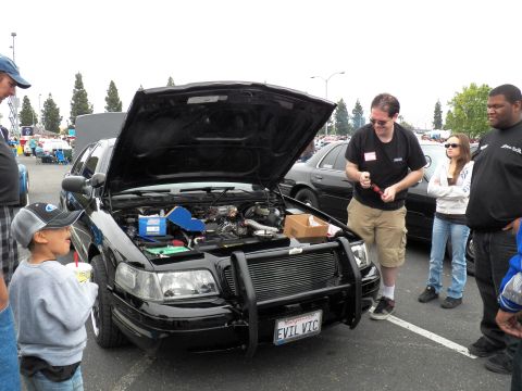 2003 Ford Crown Victoria - Spectators including a child gather around a black sedan with grille guard and EVIL VIC plate.