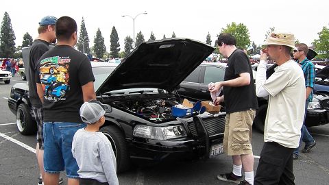 2003 Ford Crown Victoria - group converses beside the raised hood of a black sedan among rows of show cars.