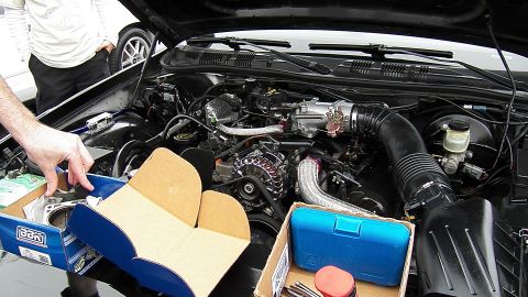 2003 Ford Crown Victoria - hand reaches into a black sedan engine bay surrounded by parts boxes and chrome intake piping.
