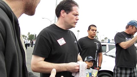 2003 Ford Crown Victoria - man in a black shirt speaks while a child and other attendees stand nearby at an outdoor meet.