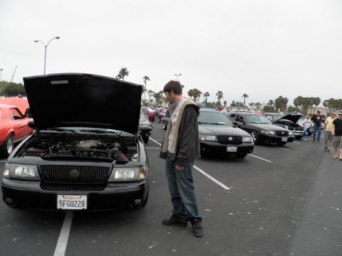 2003 Ford Crown Victoria - Man views the engine bay of a black Mercury Grand Marquis parked in a lot full of show cars under gray sky