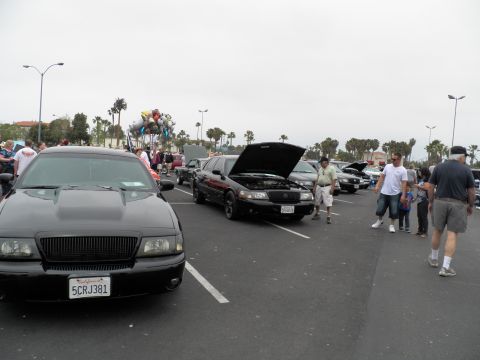 2003 Ford Crown Victoria - Attendees walk past black Mercury sedans, one with hood raised, near a cluster of balloons at an outdoor meet