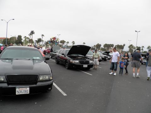 2003 Ford Crown Victoria - Spectators and children wander a car show aisle past black Mercury sedans, one with its hood propped open