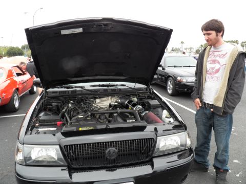 2003 Ford Crown Victoria - young man in a graphic tee stands beside a black Mercury sedan displaying its modified engine bay
