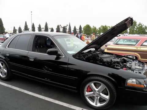 2003 Ford Crown Victoria - Profile of a black Mercury Marauder sedan with hood raised at a car show beside a wood-paneled wagon