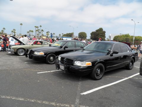 2003 Ford Crown Victoria - Black Crown Vic with front push bar and California plate, beside a green sedan and crowd of spectators