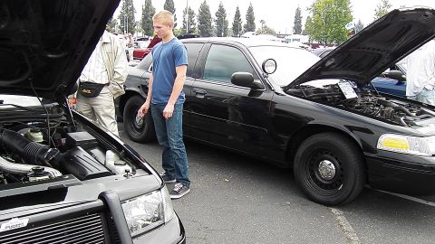 2003 Ford Crown Victoria - Young man in blue stands between two black Crown Vics with hoods raised, engine bays exposed