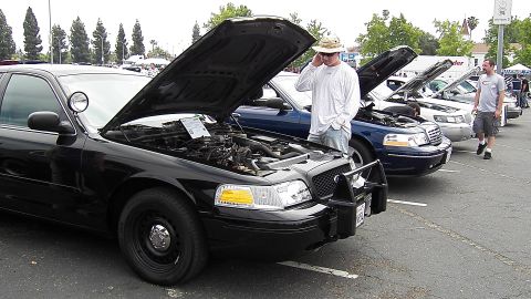 2003 Ford Crown Victoria - Lineup of black Crown Vics with raised hoods; man in straw hat inspects engine, push bar in foreground