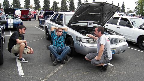 2003 Ford Crown Victoria - Three men crouch and sit beside a silver Mercury Grand Marquis with hood raised in a packed show lot