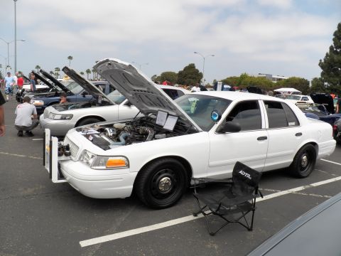 2003 Ford Crown Victoria - White Crown Vic, hood raised showing engine, steel wheels and a camp chair beside it in a busy lot