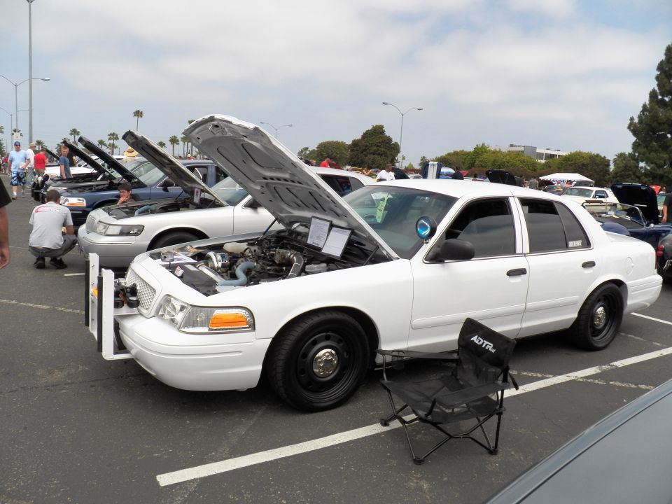 White Ford Crown Victoria with open hood and folding chair at outdoor car show