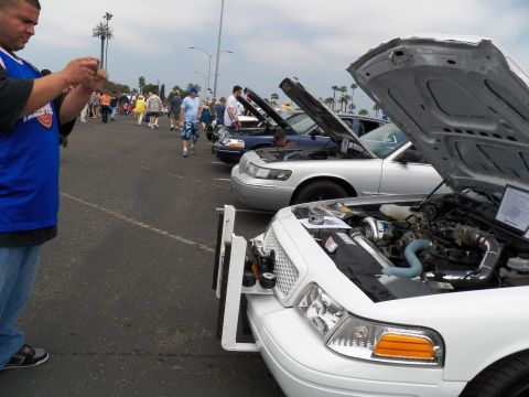 2003 Ford Crown Victoria - Man photographs a white Crown Vic with raised hood and chrome intake piping; sedans line the lot