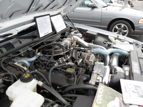 2003 Ford Crown Victoria - Engine bay of a Ford sedan showing a belt-driven blower and intake plumbing, two spec cards propped upright, COUCH banner at lower right.