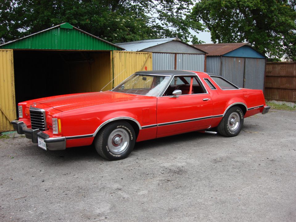 Red 1977 Ford Thunderbird coupe with a black vinyl roof parked in front of garages