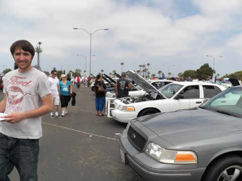 2003 Ford Crown Victoria - Smiling man in a Ford graphic tee at left, white Panther sedans and a gray Ford front in a coastal parking-lot car show behind him.