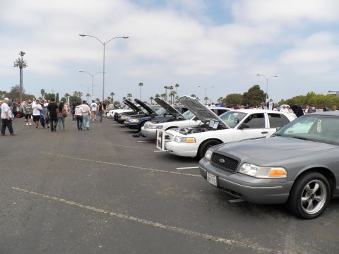 2003 Ford Crown Victoria - Line of Panther-platform Fords with open hoods, gray Crown Victoria in front, attendees and a flagpole tower in the background.
