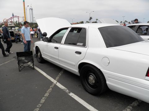 2003 Ford Crown Victoria - White Panther sedan on black steel wheels, hood and trunk raised, a man inspecting it; roller coaster visible behind the lot.