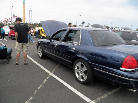 2003 Ford Crown Victoria - Blue Crown Victoria displayed hood-up; spectator in black tee stands at left near amusement park