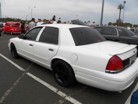 2003 Ford Crown Victoria - White Crown Victoria with trunk spoiler shown beside a red hot rod under cloudy sky