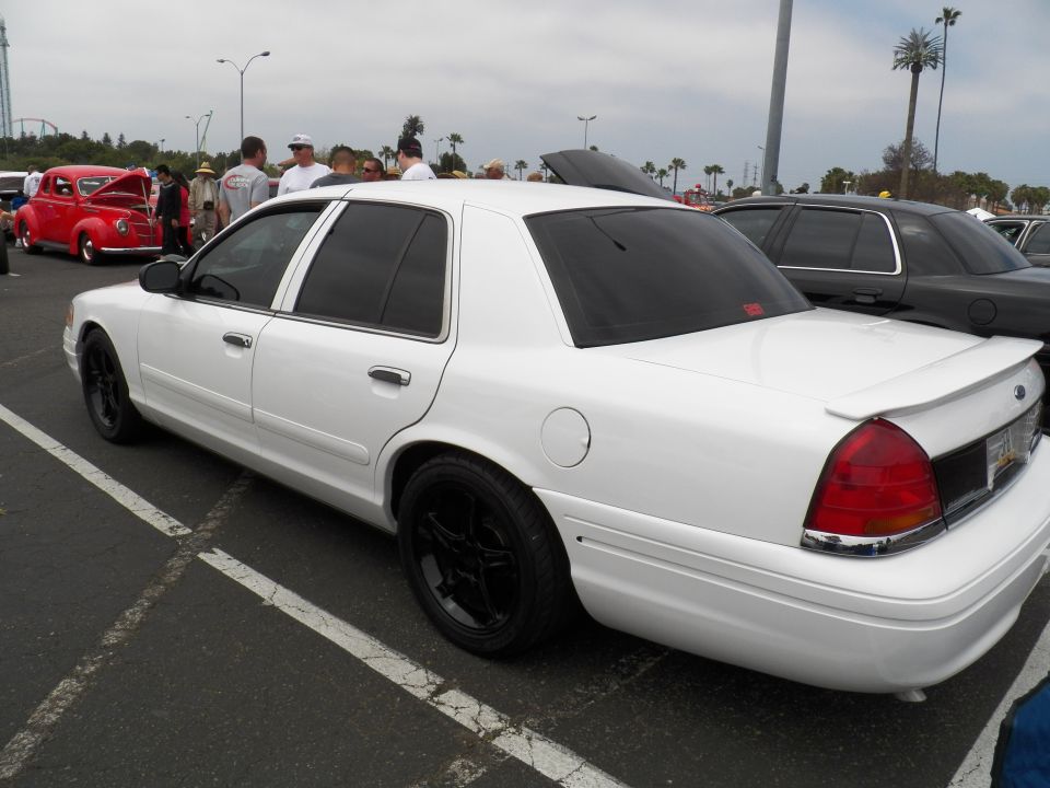 White Ford Crown Victoria sedan with rear spoiler and black wheels, red hot rod parked behind