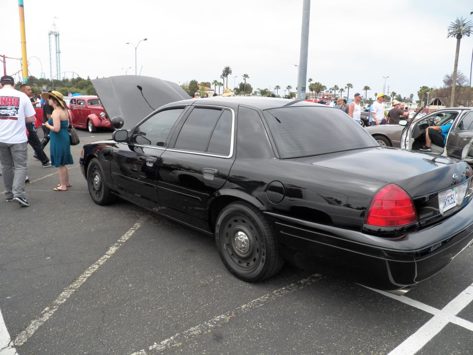 Black Ford Crown Victoria sedan with open hood and black steel wheels at car show, people nearby