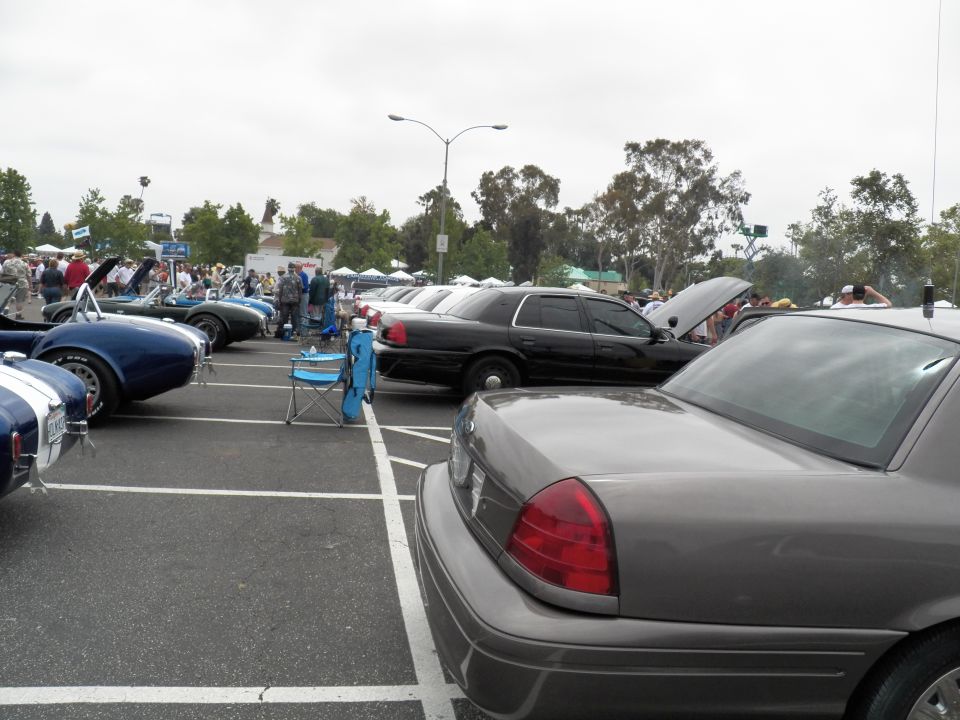 Row of sedans and a blue Shelby Cobra replica parked at outdoor car show under gray sky