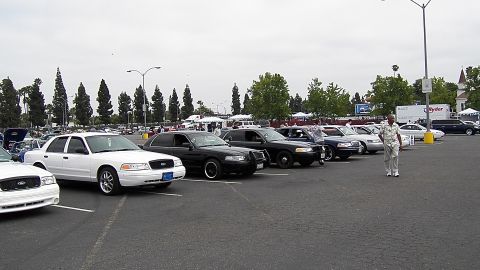2003 Ford Crown Victoria - man in fatigues stands beside a long row of Crown Victoria police sedans under a gray sky