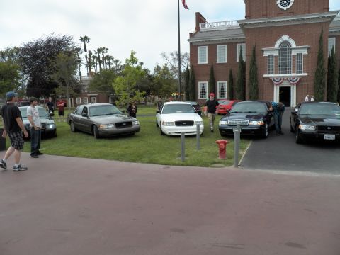 2003 Ford Crown Victoria - Onlookers walk past gray, white and black Crown Victoria sedans on a lawn by a brick building