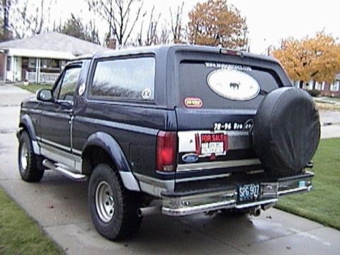 1995 Ford Bronco - Rear three-quarter view of a dark blue two-tone full-size Ford Bronco parked in a suburban driveway with autumn trees behind it. A black-co…