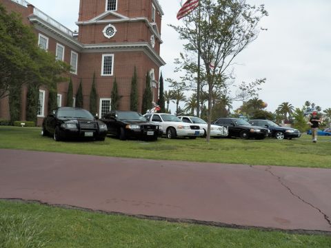 2003 Ford Crown Victoria - man walks past Crown Victoria sedans parked on grass by a brick building flying a flag