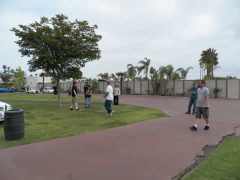 2003 Ford Crown Victoria - Several people gather near a barrel on a lawn beside a maroon path, palms and wall behind.