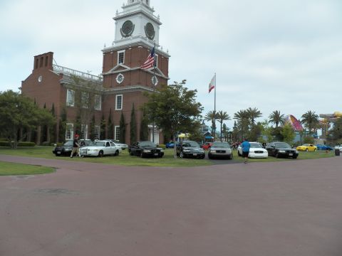 2003 Ford Crown Victoria - Mixed black and white sedans on grass before a tall white-towered brick hall under gray sky.