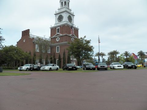 2003 Ford Crown Victoria - Long lineup of sedans parked on grass before a brick clock-tower building and palm trees.