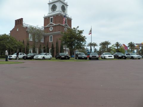 2003 Ford Crown Victoria - Mixed sedans parked across the grass before a tall white-towered brick building under gray sky.